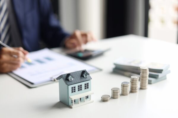 Na stolu se nalazi maketa kuće i naslagani novčići dok osoba u pozadini računa troškove i porez na izdavanje nekretnine