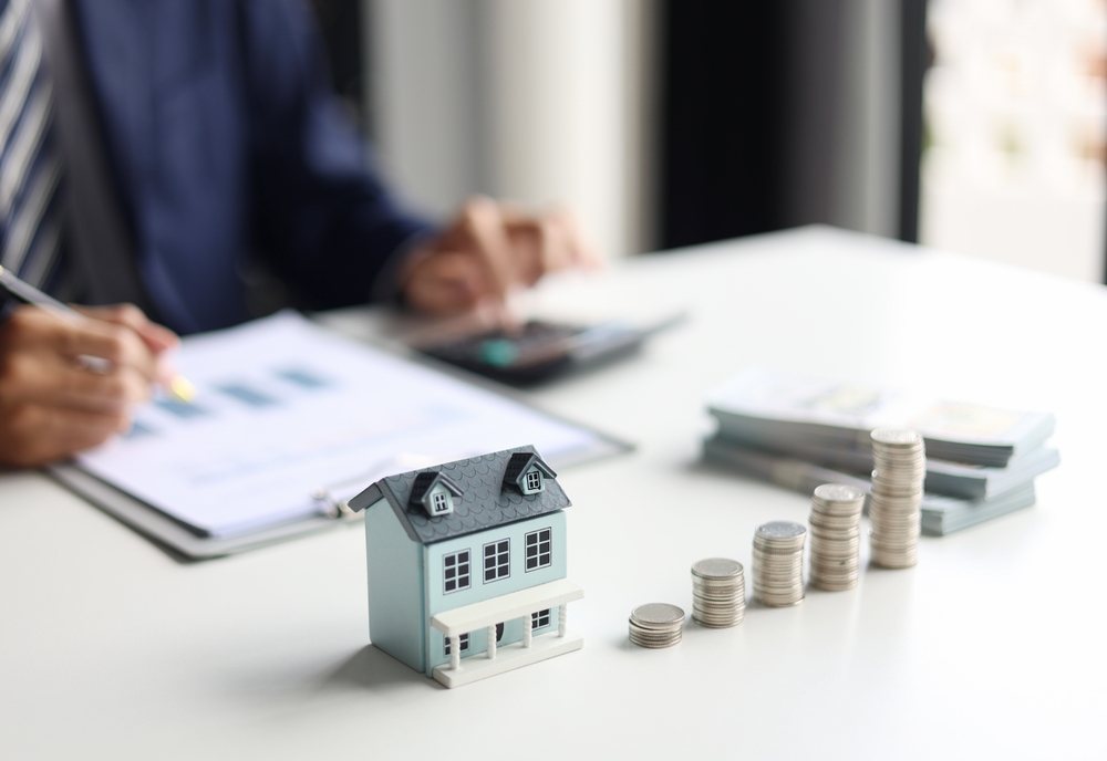 Na stolu se nalazi maketa kuće i naslagani novčići dok osoba u pozadini računa troškove i porez na izdavanje nekretnine