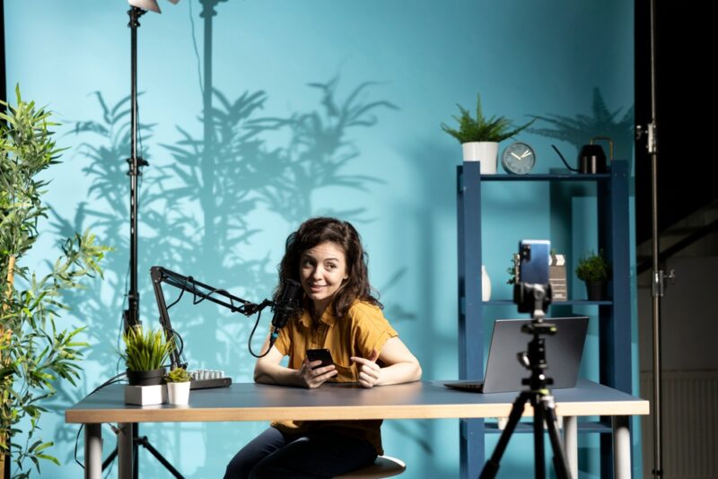 Mlada žena snima sadržaj za društvene mreže u kućnom studiju koristeći mikrofon i telefon tokom influenser kampanje.